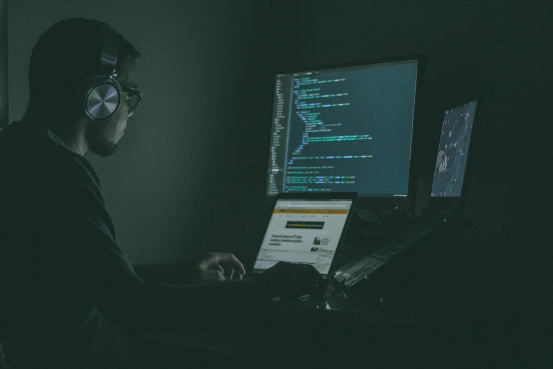 A person in headphones working at a laptop and monitors in a dark room
